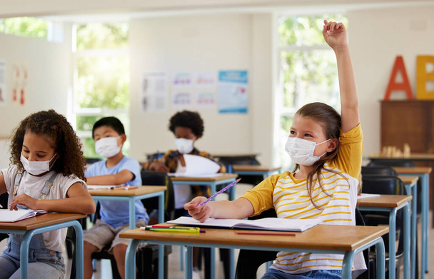 Wearing face mask to protect from covid while learning in class, answering education question and studying with students in a classroom. Girl sitting at a desk and raising hand during a pandemic. - Foto, imagen