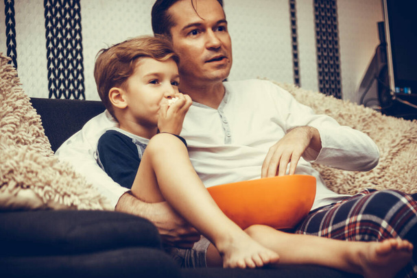 Обнятые отец и сын смотрят телевизор, расслабляясь на диване и поедая попкорн.  - Фото, изображение
