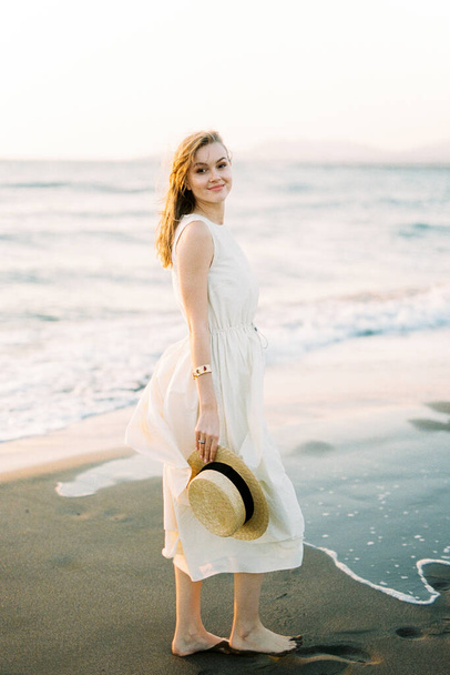 Smiling woman in a dress with a straw hat in her hand stands on the sand near the sea. High quality photo - Foto, Bild