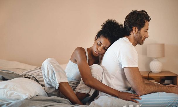 Sad, frustrated and unhappy man and woman hugging after a breakup in their bedroom. Upset, annoyed and depressed male getting support with his wife. Interracial couple cuddling to comfort husband - 写真・画像