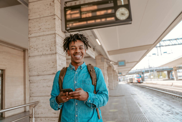 Travel. Dark-skinned cheerful guy with smartphone smiling at camera standing outdoors near railway on fine day - Photo, image