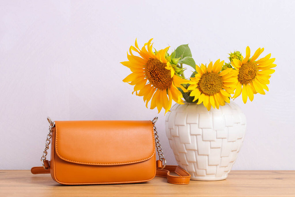 Orange women's crossbody bag and sunflowers in vase on wooden background. - Photo, image