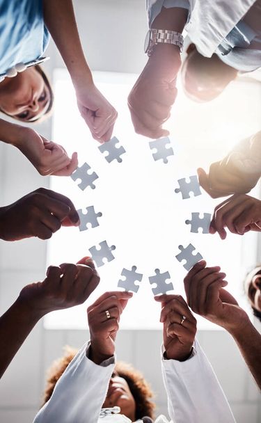 Their circle stands strong. Low angle shot of a group of doctors forming a huddle while each holds a puzzle piece inside of a hospital - Foto, Imagen