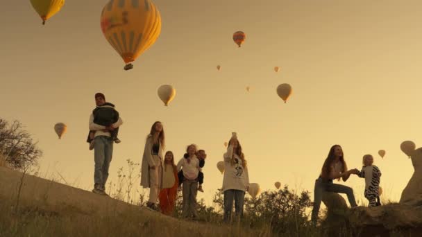 Low angle of group of friends or two families of parents with children watching and admiring colorful hot air balloons flying in cloudless sky at sunrise time in valley during trip in Cappadocia. 4k - Séquence, vidéo