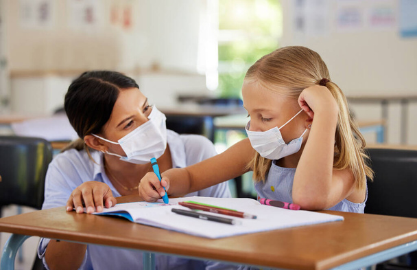 Education, covid and learning with face mask on girl doing school work in classroom, teacher helping student while writing in class. Elementary child wearing protection to stop the spread of a virus. - Photo, image