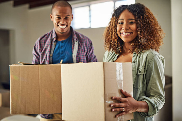 Our life is beginning one step at a time. Portrait of a cheerful young couple carrying boxes together to move into their new home inside during the day - Foto, imagen
