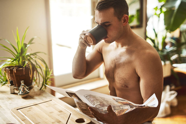 Τίποτα καλύτερο από ένα φλιτζάνι καφέ. Ένας όμορφος νέος χωρίς πουκάμισο πίνει καφέ και διαβάζει εφημερίδα στην κουζίνα στο σπίτι. - Φωτογραφία, εικόνα
