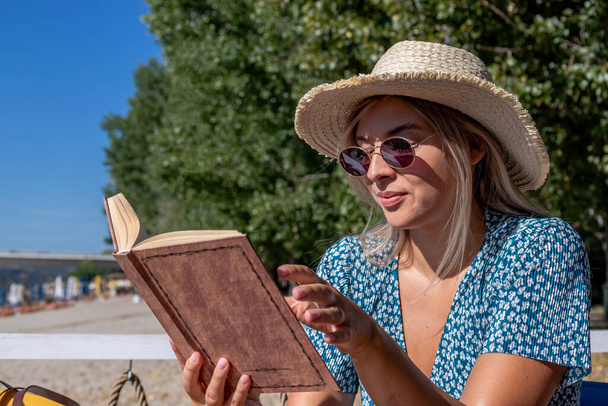 A beautiful young woman wearing a straw hat and sunglasses, reading a book and enjoying a lovely summer morning in a beach cafe by the river. - Foto, Imagem