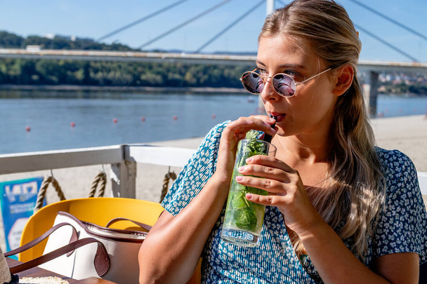 A beautiful blonde woman with sunglasses is having a cocktail and enjoying a lovely summer afternoon in a beach cafe by the river. - Fotó, kép