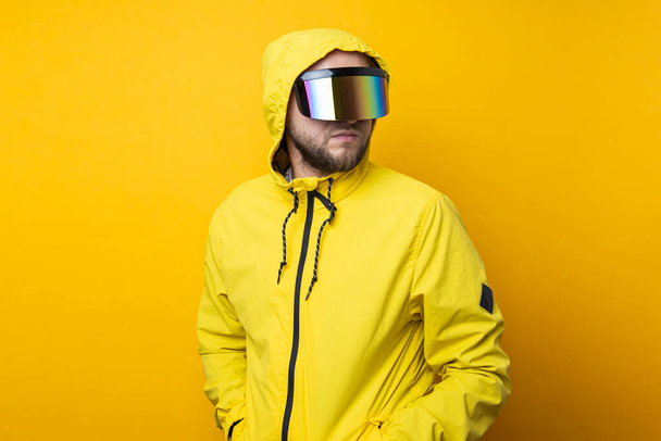 Νεαρός με γυαλιά εικονικής πραγματικότητας με κίτρινο μπουφάν κοιτάζει μακριά σε κίτρινο φόντο. - Φωτογραφία, εικόνα