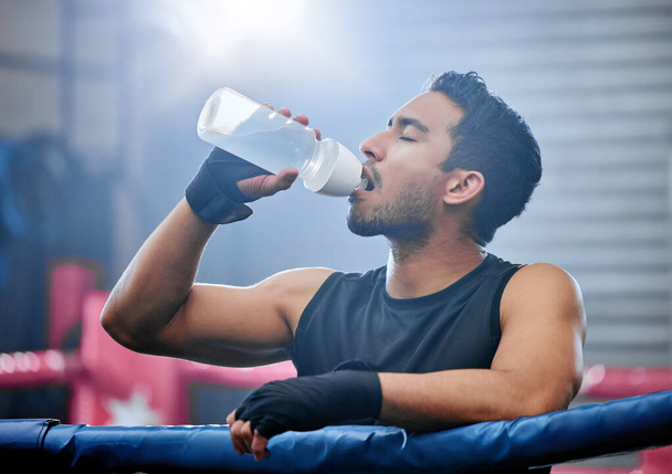 Fit, active and healthy boxer drinking water, on break and staying hydrated in routine workout, training or boxing ring exercise. Sporty, athletic or strong man after kickboxing fight or sports match. - 写真・画像