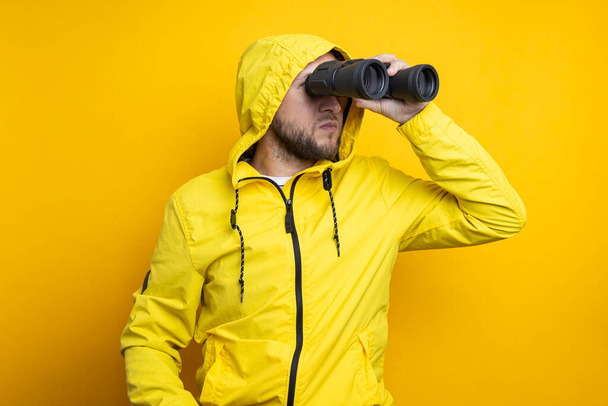 Young man in yellow raincoat looking through binoculars on yellow background. - Fotó, kép