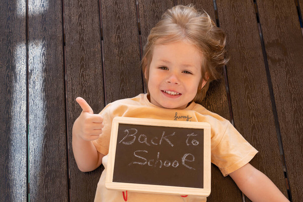 Happy caucasian child boy 3 years old showing a blackboard with smile lying on a table in the terrace outdoors.Back to school concept. - Foto, afbeelding