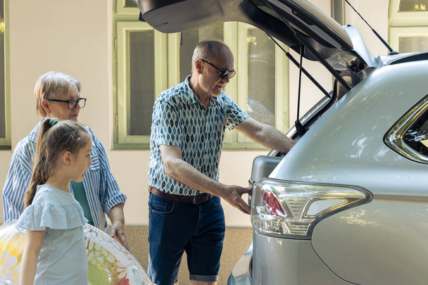 Senior people taking child on holiday by car, travelling on summer vacation at seaside. Grandparents and niece leaving on journey adventure, putting travel bags and inflatable in trunk. - Zdjęcie, obraz