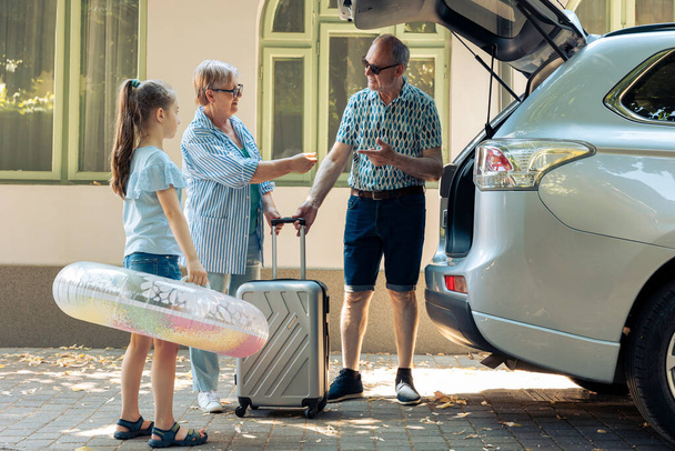 Grandparents and niece leaving on holiday, travelling on seaside vacation during summer. Senior couple and little girl going on journey trip with travel bags and inflatable in trunk of vehicle. - Photo, image