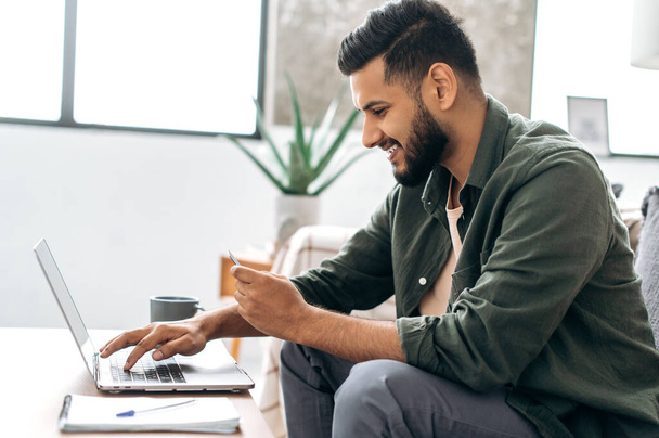 Side view of satisfied positive indian or arabian guy in stylish clothes sitting on a sofa in a living room, using laptop and bank card to pay online, ordering goods and clothes, home delivery, smiles - Foto, Imagen