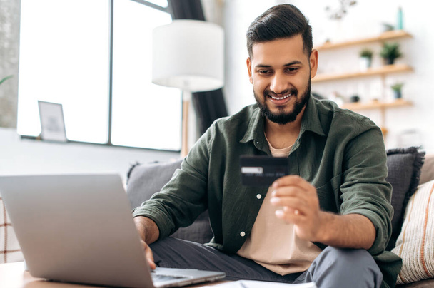 Online payment. Indian or Arabian positive young male in casual wear, sits in cozy living room, holding bank card in hand, using laptop, shopping online, ordering home delivery, paying bills, smiling - Φωτογραφία, εικόνα