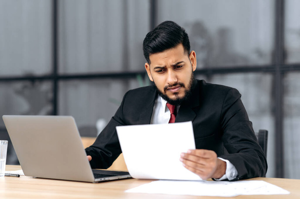 Разочарованный уставший индийский или арабский бизнесмен, расстраивающийся за рабочим столом с ноутбуком в современном офисе, работающий с документами, составляющий финансовый отчет, анализирующий результат работы, чувствует себя перегруженным работой - Фото, изображение