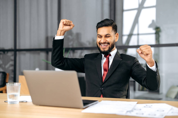 Excited indian or arabian businessman in suit, sits in a modern office, looking at laptop, happy by good news, rejoice by win, raising hand in yes gesture, celebrating project success, big profit - Foto, imagen