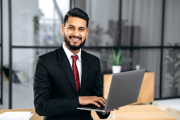 Портрет успешного уверенного элегантного индийского или арабского молодого бизнесмена, в костюме, держащего открытый ноутбук в руке, стоящего рядом с рабочим столом в своем современном офисе, смотрящего на камеру, улыбающегося - Фото, изображение
