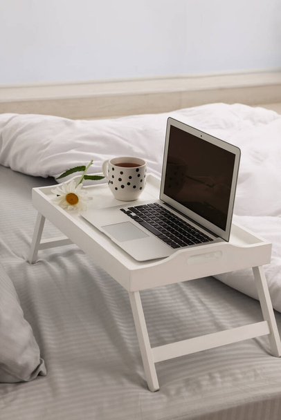 White tray table with laptop, cup of drink and daisy on bed indoors - Foto, Bild