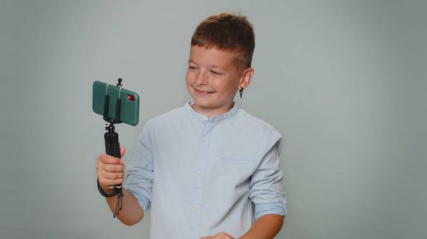 Toddler boy kid traveler blogger taking selfie on mobile phone selfie stick communicating video call online with subscribers. Young children teenager. Child isolated alone on gray studio background - Photo, image