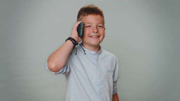 Toddler kid boy in shirt having pleasant mobile conversation using smartphone with friends or family, telling good news. Young teenager child schoolboy isolated alone on gray studio background indoors - Photo, Image