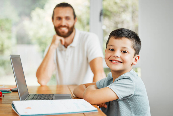 Μαθαίνοντας, διδάσκοντας και χαμογελώντας μικρό αγόρι κάνει σπίτι σχολική εργασία σε απευθείας σύνδεση σε ένα φορητό υπολογιστή. Ευτυχισμένος γιος με στοργικό πατέρα στο παρασκήνιο έτοιμος να μάθει, να μελετήσει και να διασκεδάσει ψηφιακά στο σπίτι της οικογένειας. - Φωτογραφία, εικόνα