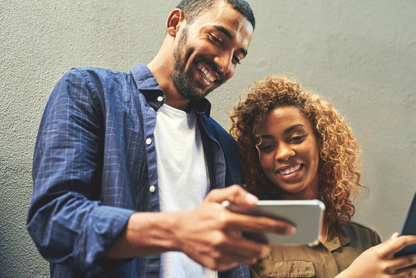 Smiling young couple looking, laughing and holding phone together showing a funny social media app. Friends sharing a gossip news article post online. Guy looking at internet web content with girl. - Foto, imagen