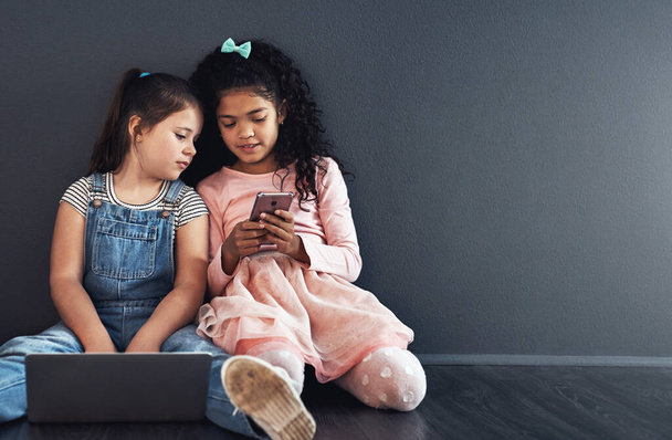 Родился в поколении экрана. Студийный снимок двух молодых девушек, сидящих на полу и использующих беспроводные технологии на заднем плане - Фото, изображение