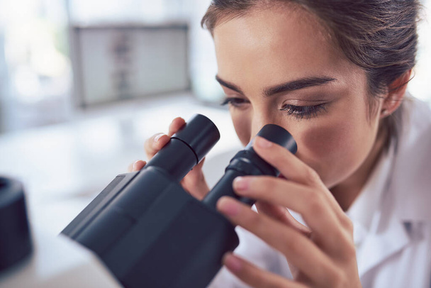 A detailed point of view. Closeup of a focused young female scientist looking through the lens of a microscope inside of a laboratory - Photo, Image