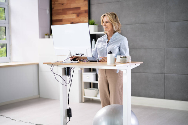 Woman Using Adjustable Height Standing Desk In Office For Good Posture - 写真・画像