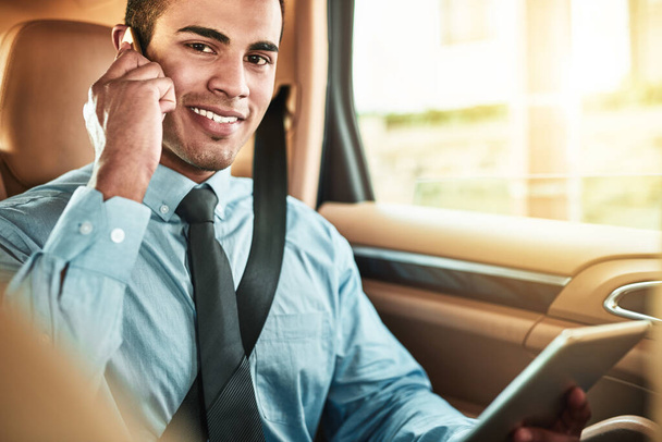 Hes the executive everyone wants to get in touch with. Portrait of a young businessman talking on a phone while using a digital tablet in the back seat of a car - Photo, Image