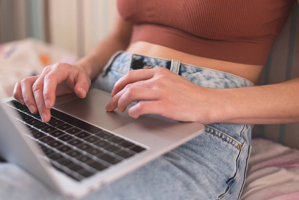 Una chica trabaja desde casa, las manos femeninas en un teclado portátil de cerca, una mujer se sienta en un sofá con un ordenador portátil - Foto, Imagen