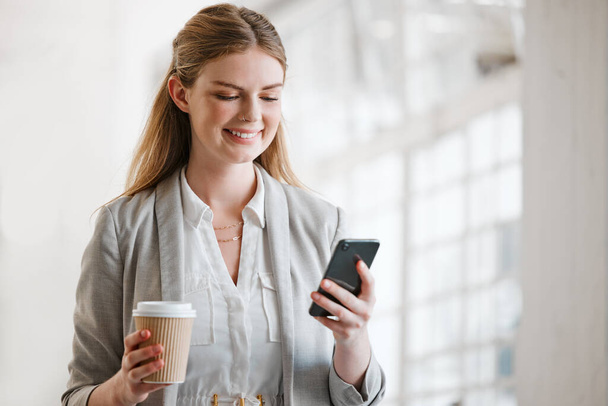Phone, coffee and online of a young employee texting on smartphone in a corporate office. Happy, smile and mobile woman on the internet at work networking, news and social media at the office - Photo, image