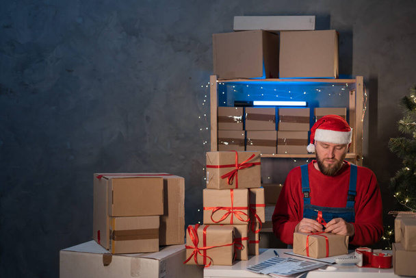 bearded man wearing a Santa Claus hat works in a warehouse of boxes with gifts and orders from an online store for Christmas. Small Business Owner Sells Happy New Year. Delivery holiday. - Fotoğraf, Görsel