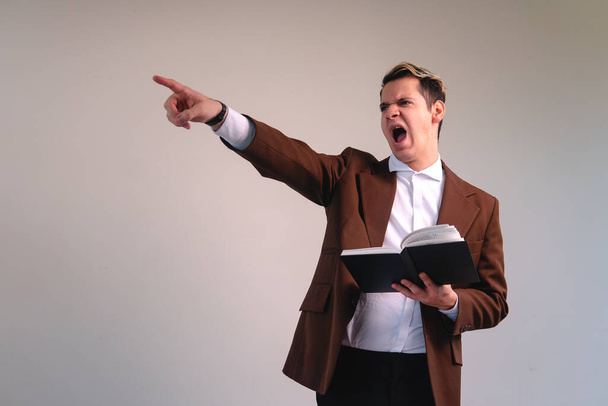 Caucasian blond man dressed in a suit and with a book in his hand. The man is shouting euphorically and pointing somewhere. Annoyed lawyer pointing. Very expressive teacher. High quality photo - 写真・画像