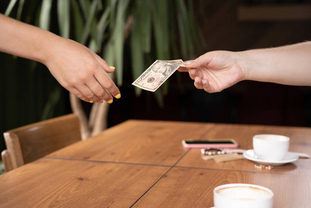 Γυναίκα χέρι κρατώντας χαρτονομίσματα των ΗΠΑ δίνοντας σερβιτόρα καφέ, δύο φλιτζάνια καφέ στο παρασκήνιο. πληρωμή, χρέωση υπηρεσιών, έλεγχος λογαριασμού, ιδέες συμβουλές χρημάτων - Φωτογραφία, εικόνα