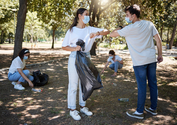 Ομίχλη, καθαρισμός και εθελοντές που φορούν μάσκες ενώ καθαρίζουν ένα κοινοτικό πάρκο και λένε ευχαριστώ με μια χειρονομία αγκώνα. Κοινωνική αποδιοργάνωση μεταξύ φίλων που κάνουν καθαρισμό στην ύπαιθρο κατά τη διάρκεια πανδημίας. - Φωτογραφία, εικόνα