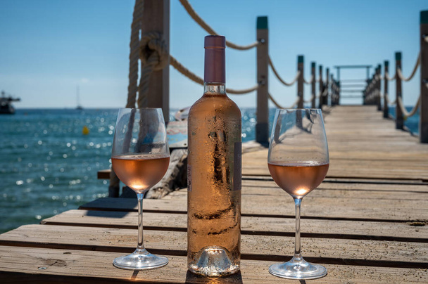 Glasses and bottle of cold rose wine from Provence served outdoor on wooden yacht pier with view on blue water and white sandy beach Plage de Pampelonne near Saint-Tropez, summer vacation in France - 写真・画像