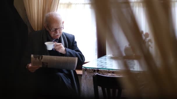 Senior man reads a newspaper at home in the kitchen, he is dressed and ready to go for a walk. High quality 4k video - Imágenes, Vídeo