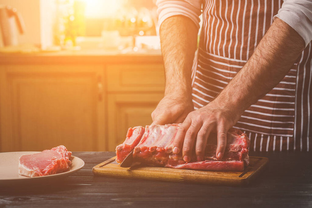 Homem cortes de carne fresca em uma tábua de corte de madeira na cozinha da casa. Um homem de avental listrado com uma faca nas mãos
 - Foto, Imagem