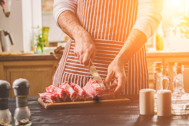 Человек режет свежую говядину на деревянной доске на домашней кухне. Мужчина в полосатом фартуке с большим ножом в руках
 - Фото, изображение