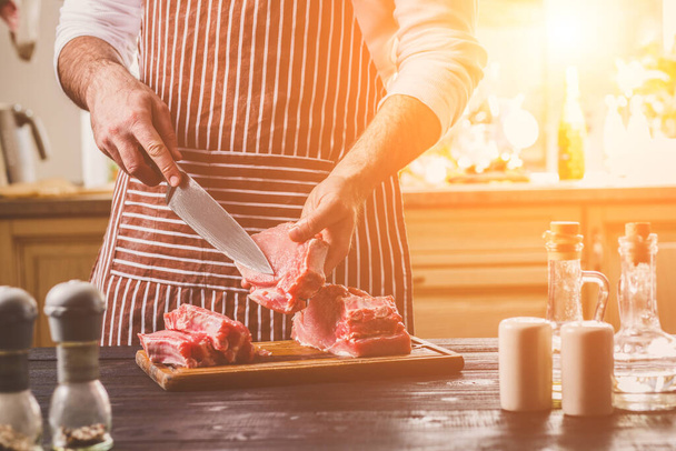 Чоловік розрізає свіжий шматок яловичини на дерев'яній обробній дошці на домашній кухні. Чоловік в смугастому фартуху з великим ножем в руках
 - Фото, зображення