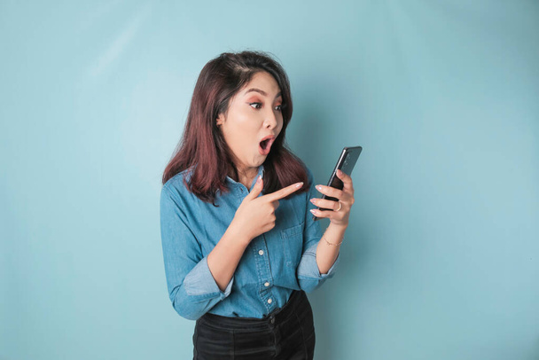 Surprised Asian woman wearing blue shirt pointing at her smartphone, isolated by blue background - Photo, image