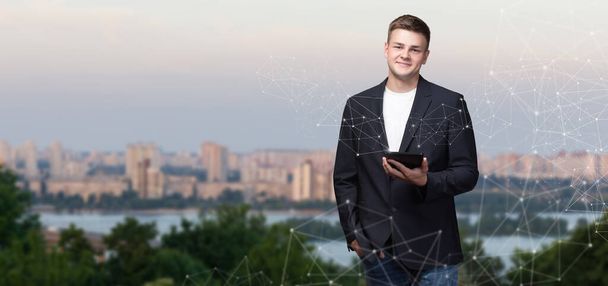 A young businessman stands with a tablet on the background of a network of connections of the city panorama. - Foto, afbeelding