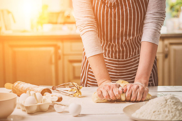 As mãos de mulher amassam a massa de farinha na mesa de cozinha. Uma mulher de avental listrado está cozinhando na cozinha
 - Foto, Imagem