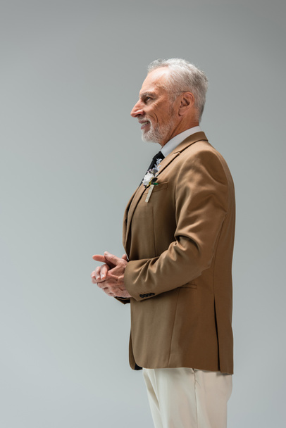 side view of happy middle aged groom in suit with floral boutonniere isolated on grey  - Foto, afbeelding