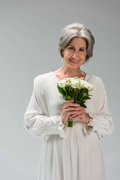 cheerful middle aged bride in white wedding dress holding bouquet isolated on grey - Photo, Image