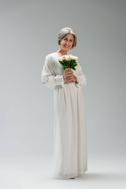 full length of happy middle aged bride in white dress holding wedding bouquet on grey - Foto, Imagen